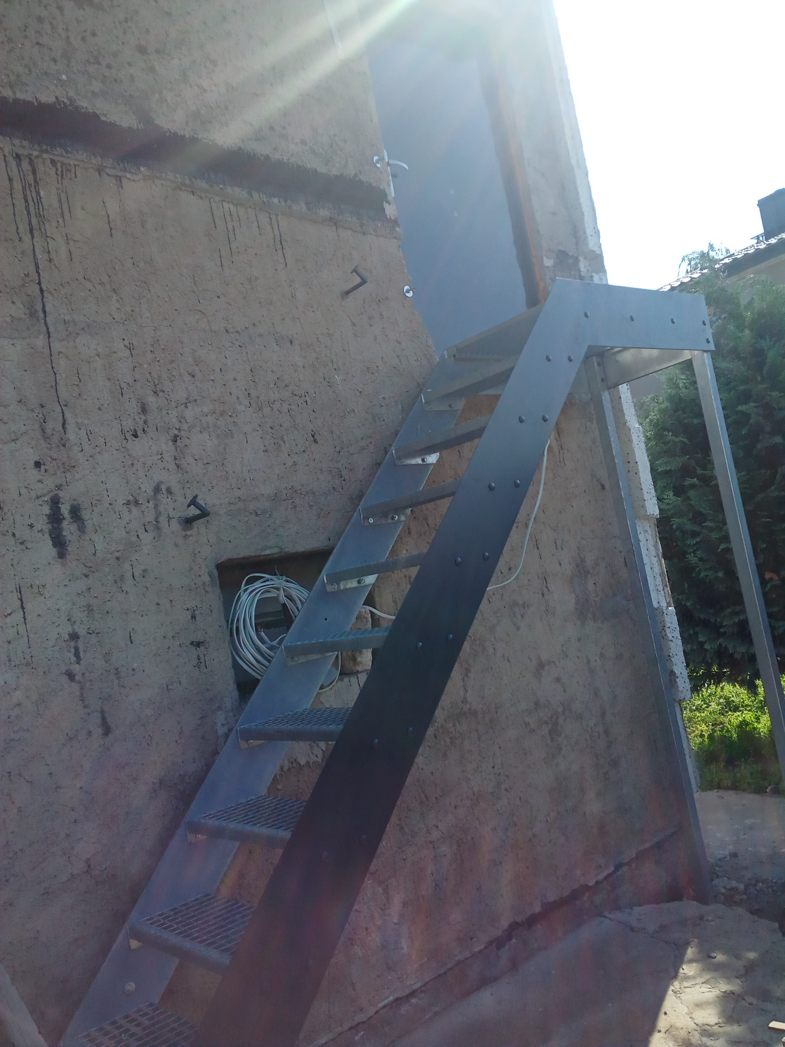 Zewnętrzne schody metalowe prowadzące do drzwi na elewacji budynku o nierównej fakturze, widoczne przewody w otworze w ścianie.