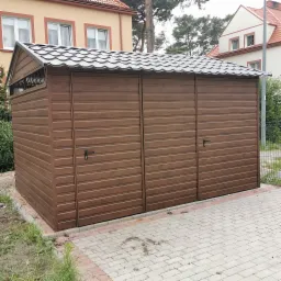 Trzysegmentowy, brązowy garaż z blachy drewnopodobnej, z szarą, falistą blachodachówką, na kostce brukowej, w tle budynki mieszkalne.
