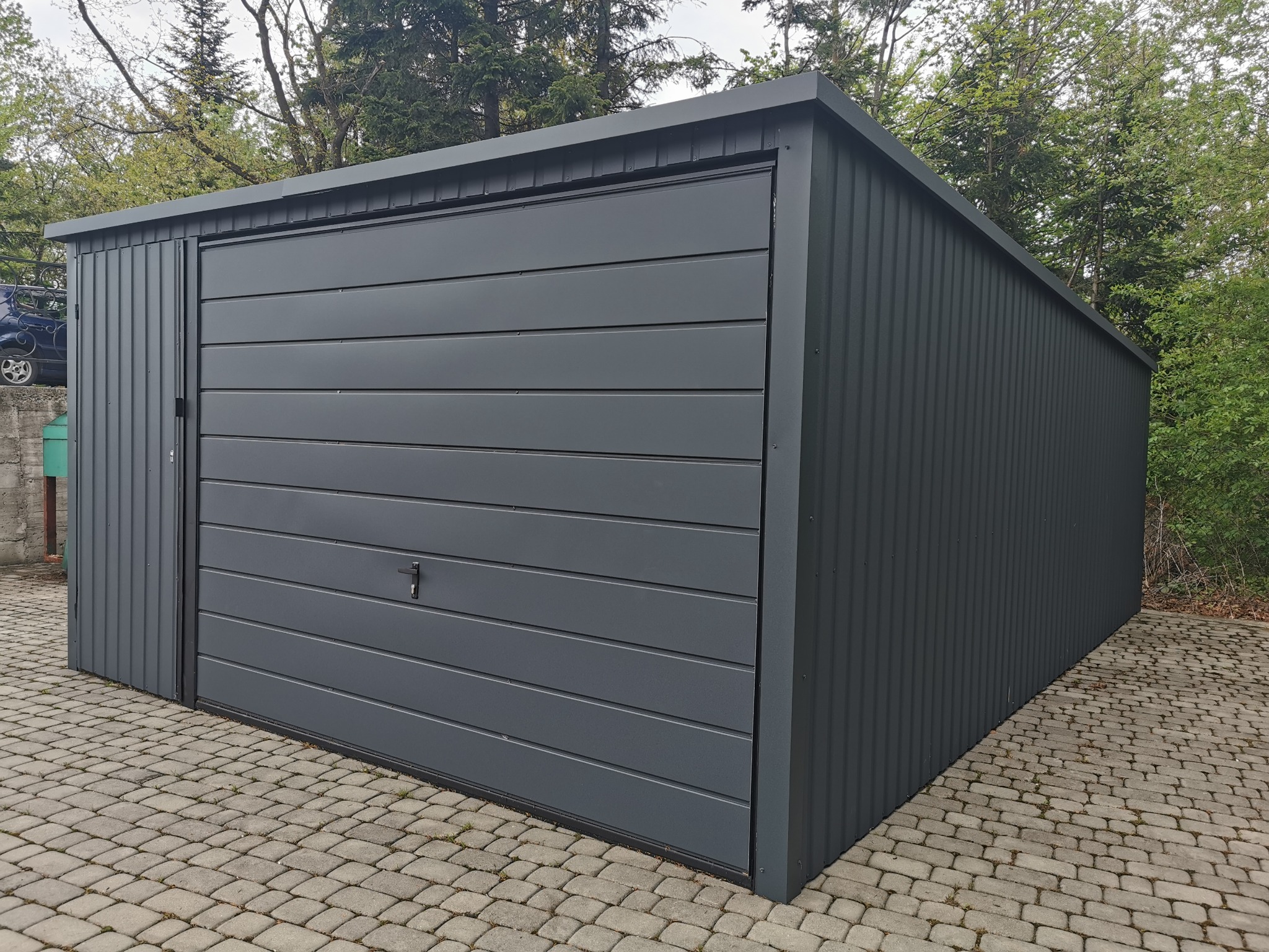 Nowoczesny, grafitowy garaż blaszany z bramą segmentową i drzwiami bocznymi, umieszczony na brukowanej powierzchni, z zielenią w tle.