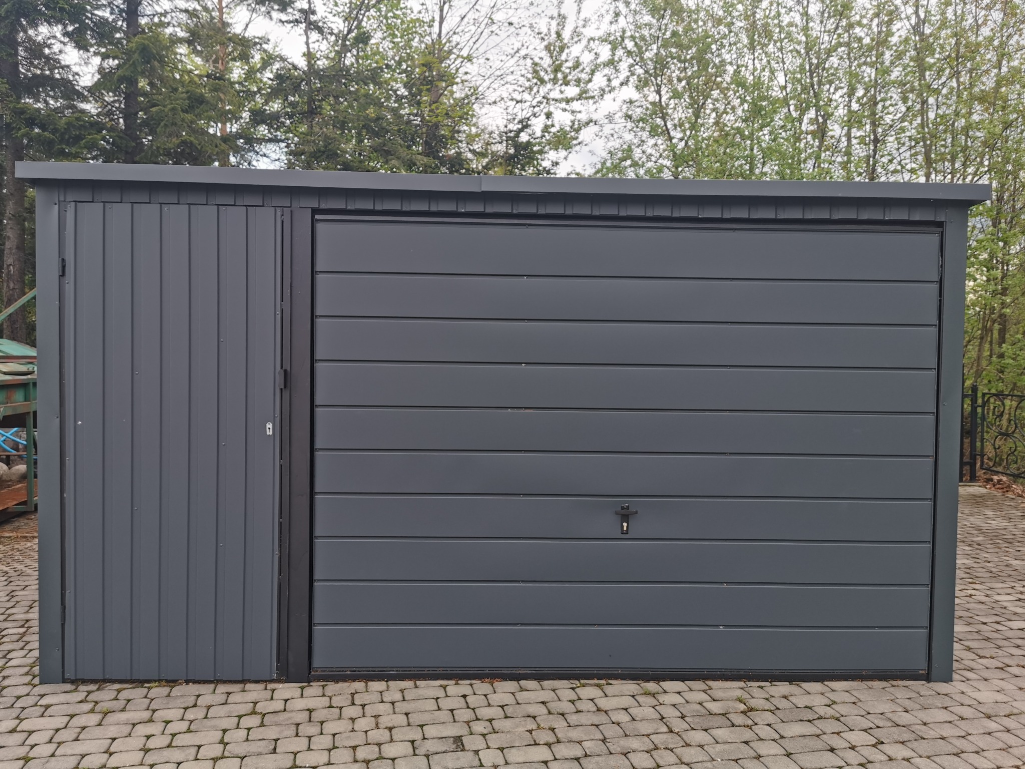 Nowoczesny, szary garaż blaszany z bocznymi drzwiami i poziomym wzorem paneli, ustawiony na kostce brukowej, z widocznymi drzewami w tle.