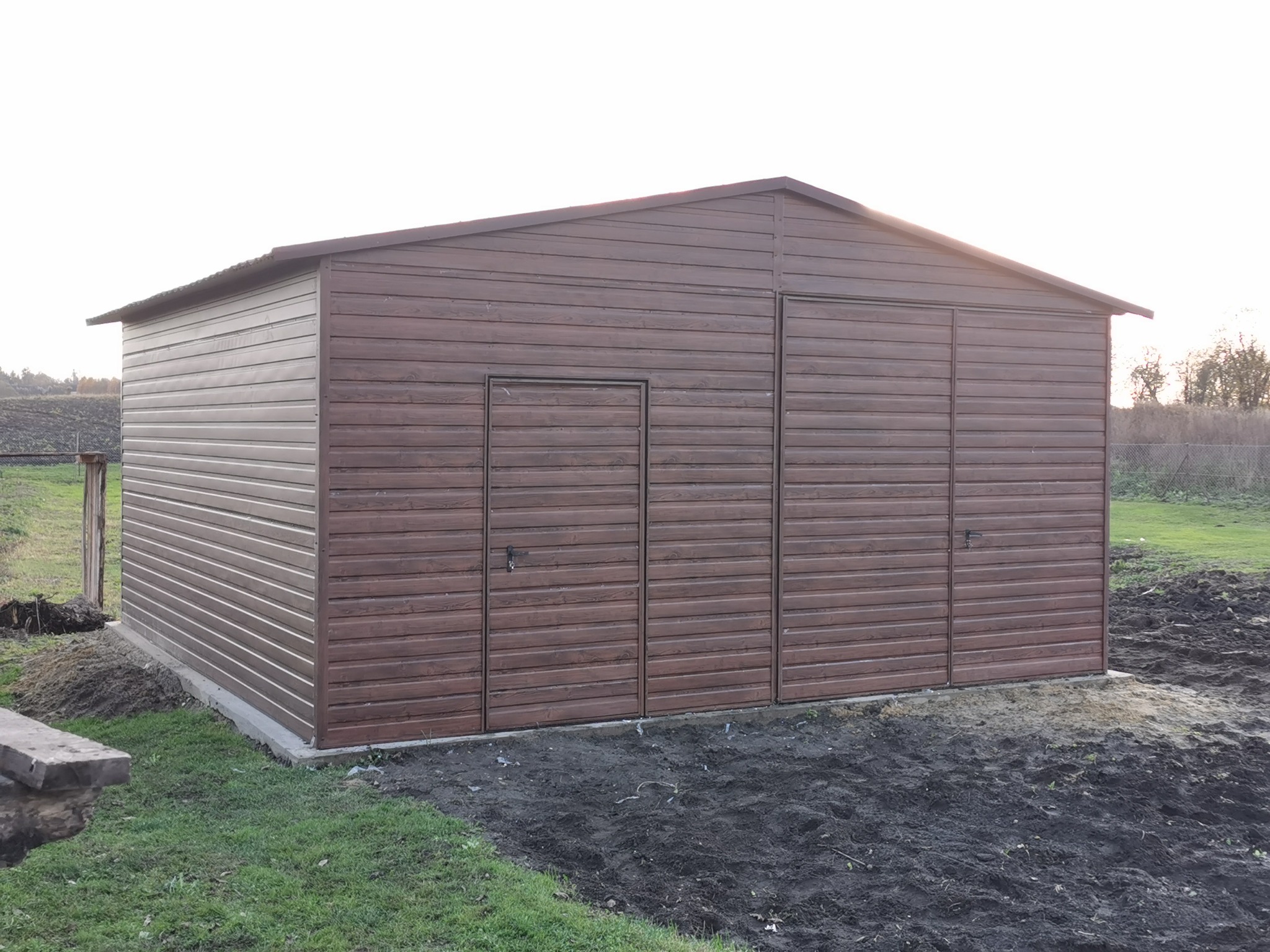 Brązowy, metalowy garaż z dwojgiem drzwi i drzwiami wejściowymi, umieszczony na betonowej podstawie na trawiastym terenie.