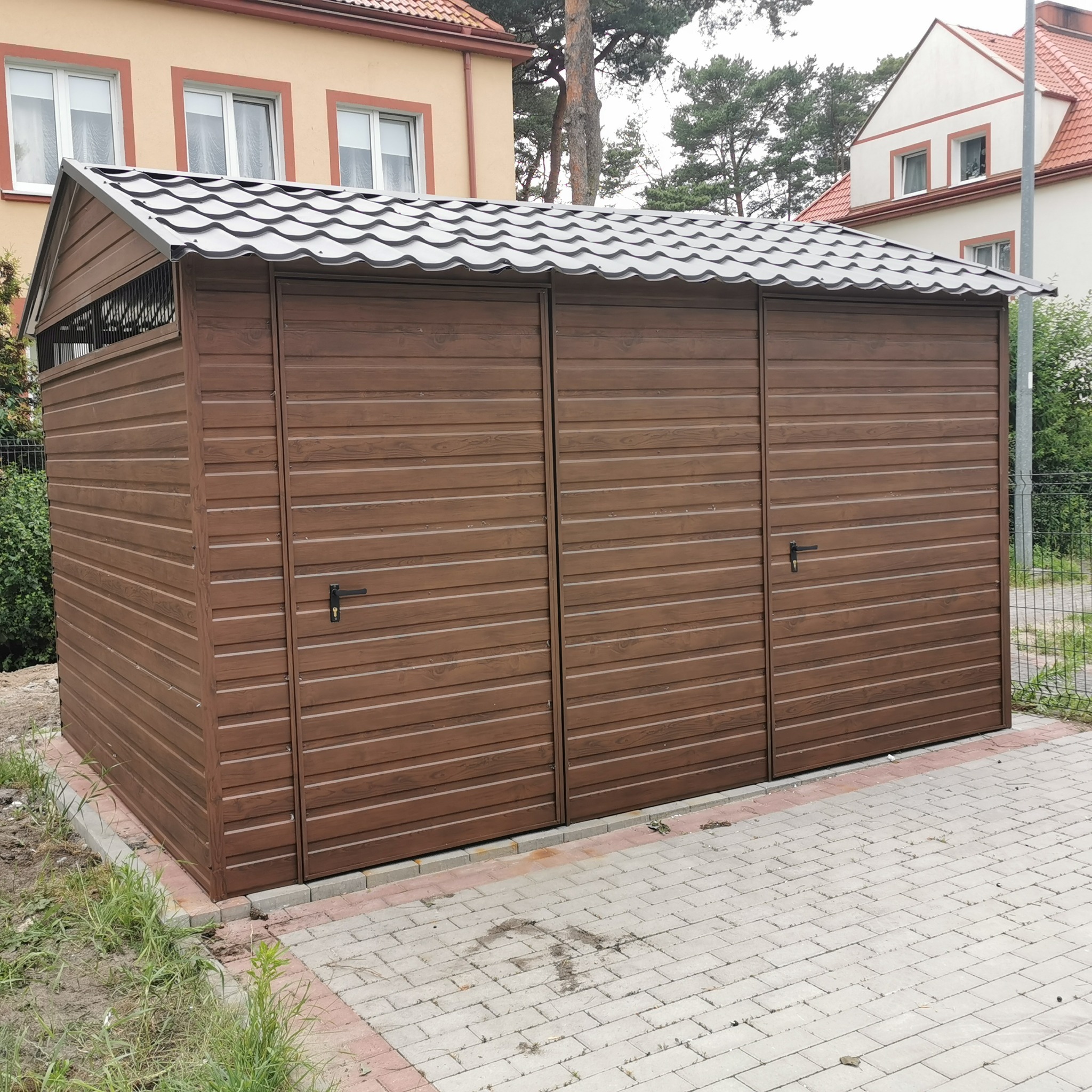 Trzysegmentowy, brązowy garaż z blachy drewnopodobnej, z szarą, falistą blachodachówką, na kostce brukowej, w tle budynki mieszkalne.