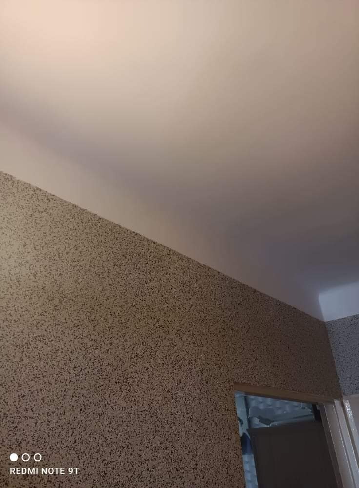 Ściana i sufit pokryte dekoracyjną strukturą, imitującą tynk mozaikowy w odcieniach beżu i brązu, widoczne z bliskiej perspektywy z fragmentem futryny drzwiowej.