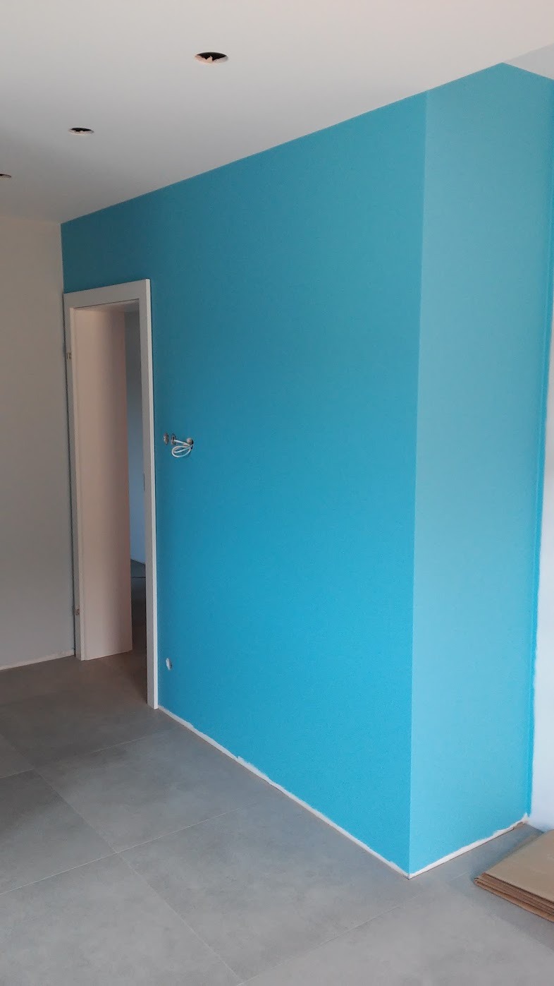 Świeżo pomalowane na turkusowo ściany w pokoju z szarą podłogą z płytek, widoczny narożnik i listwa przypodłogowa, przygotowane otwory na oświetlenie sufitowe.