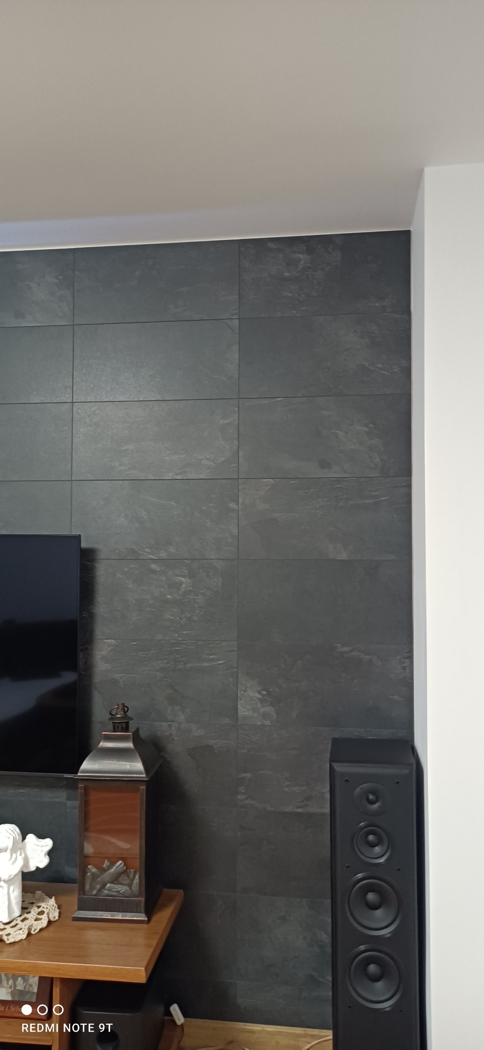 Ściana wykończona ciemnymi, kwadratowymi płytkami imitującymi kamień, obok stoi czarna kolumna głośnikowa i drewniany stolik z dekoracją, w rogu widoczna biała ściana.
