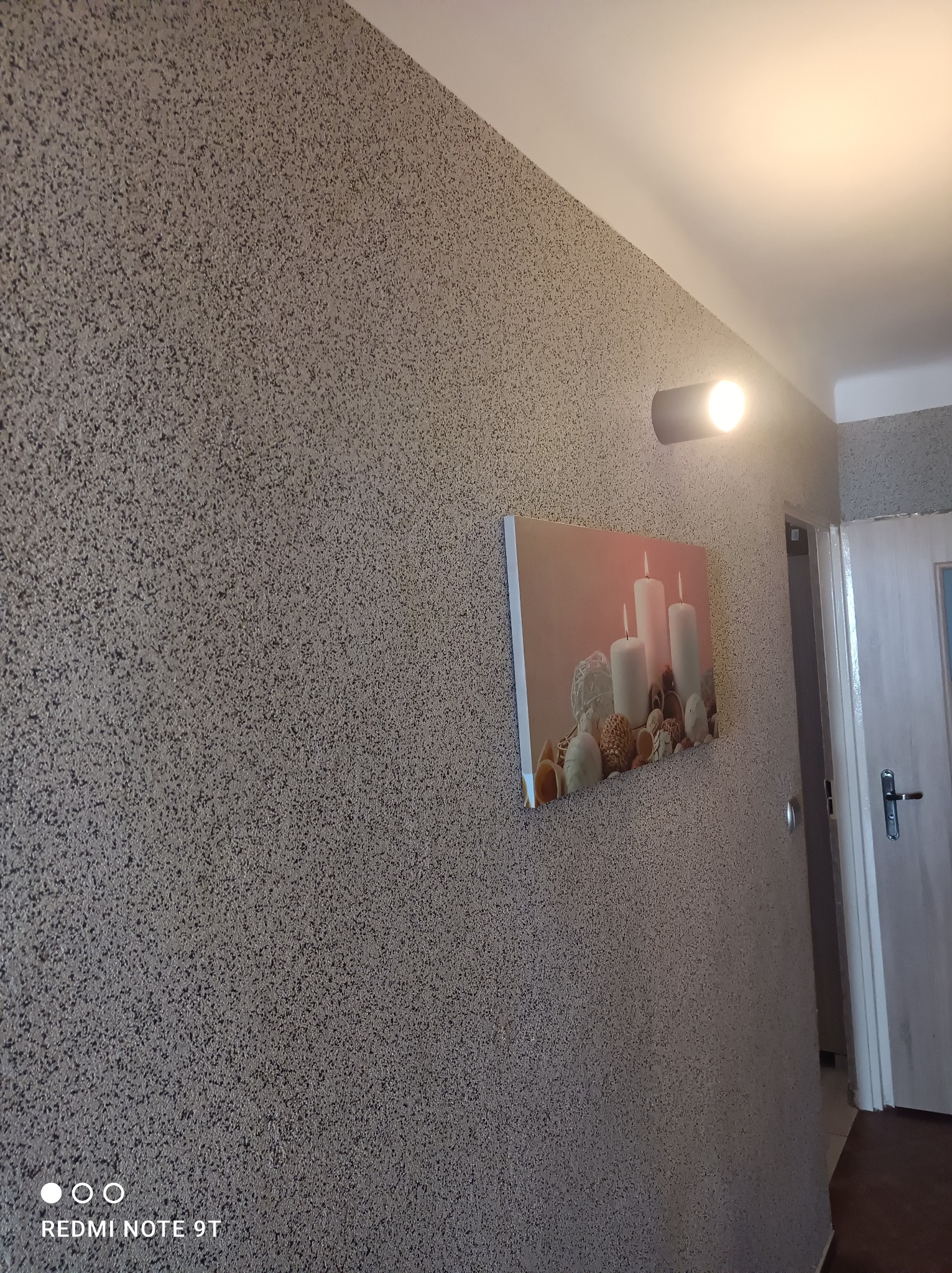 Ściana pokryta dekoracyjnym tynkiem strukturalnym w odcieniach szarości, oświetlona punktowym światłem i ozdobiona obrazem z motywem świec, widok na fragment drzwi w tle.