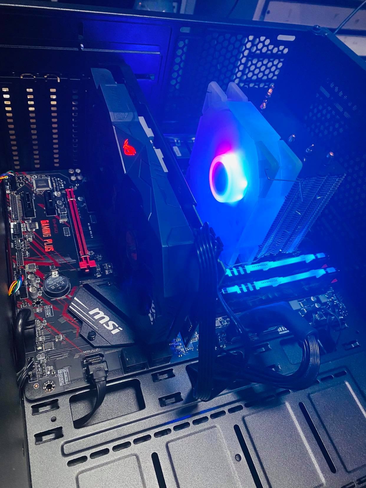 Wnętrze komputera stacjonarnego z podświetlanymi komponentami: karta graficzna ROG, płyta główna MSI Gaming Plus, chłodzenie procesora z tęczowym podświetleniem i pamięć RAM HyperX, widoczne kable...