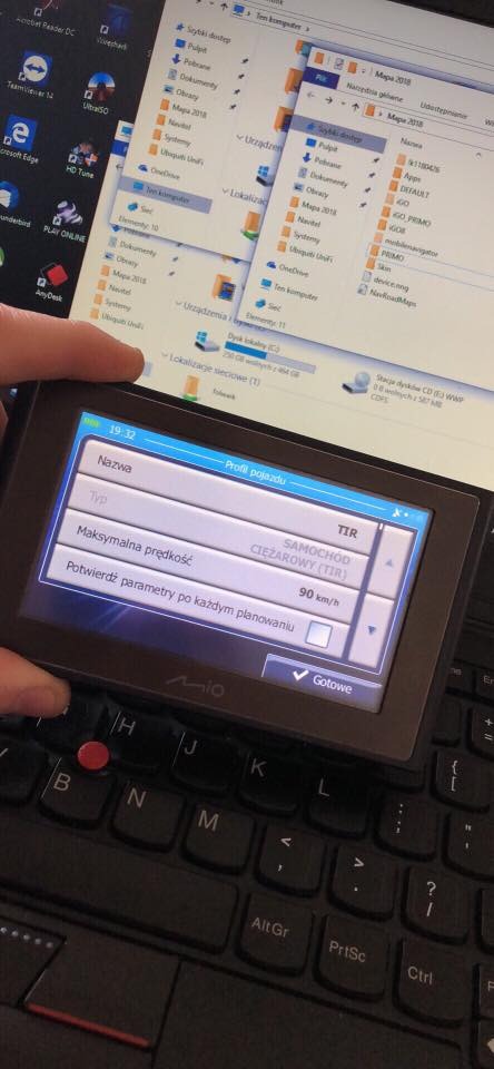 Ekran nawigacji GPS Mio leżący na klawiaturze laptopa Lenovo, wyświetlający ustawienia profilu pojazdu TIR, z widocznymi oknami eksploratora Windows w tle, sugerując konfigurację urządzenia.