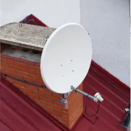 Biała antena satelitarna zamontowana na ceglanym kominie z szarą czapą, na tle czerwonego dachu i białej ściany budynku.