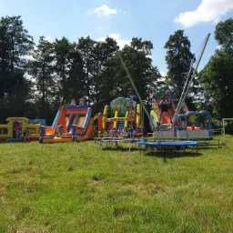 Rozległy park rozrywki na trawiastym terenie z dmuchanymi zamkami, zjeżdżalniami i trampolinami bungee dla dzieci, otoczony drzewami i błękitnym niebem.