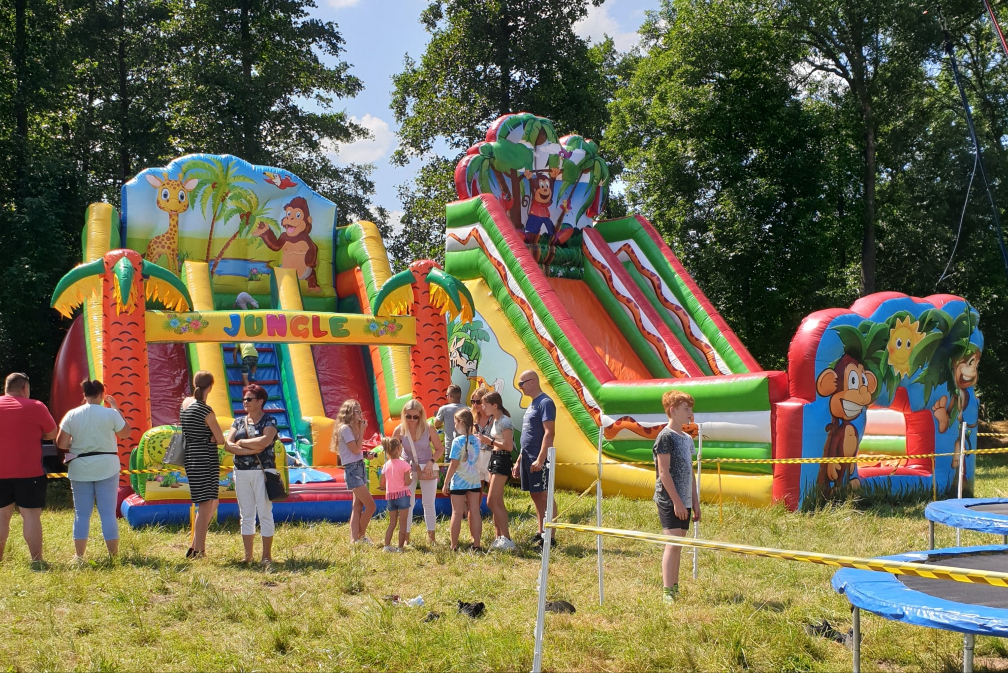 Dwa duże, kolorowe dmuchane zamki ze zjeżdżalniami, motyw dżungli, ludzie w tle, trawnik, drzewa, częściowo widoczne trampoliny.