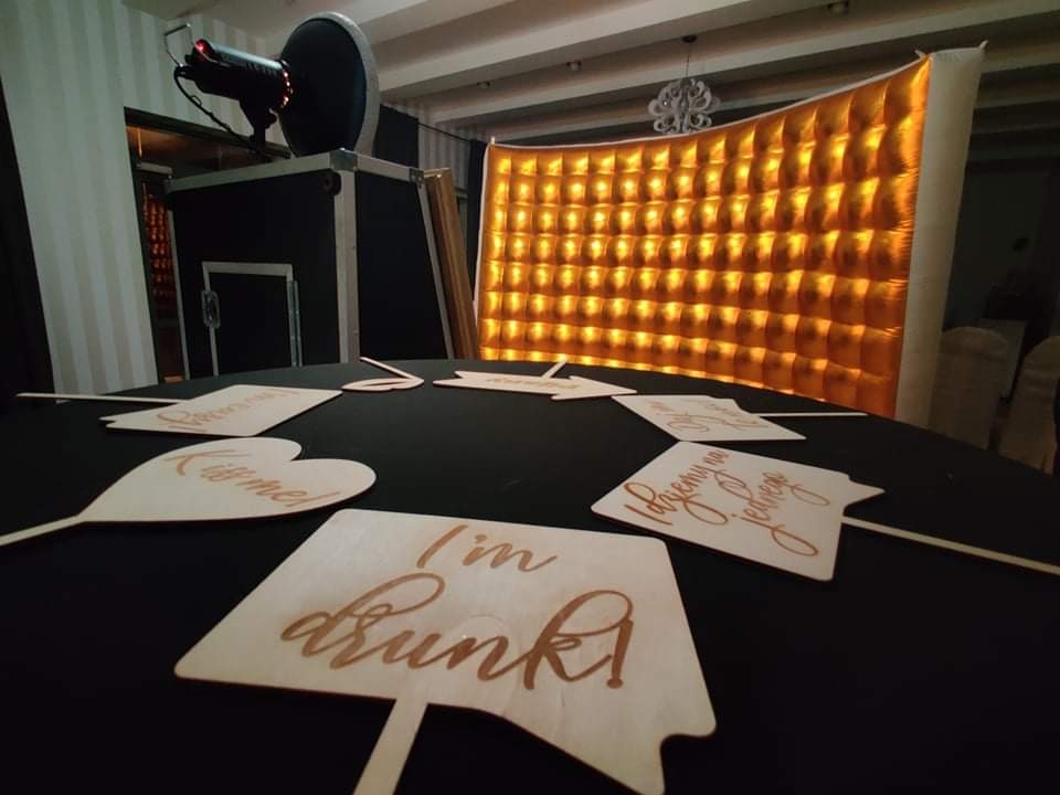 Przygotowany stół z rekwizytami do zdjęć, takimi jak tabliczki z napisami 'I'm drunk', 'Kiss me!', 'Pojemy po jednego', na tle złotej, błyszczącej ścianki i fotobudki.