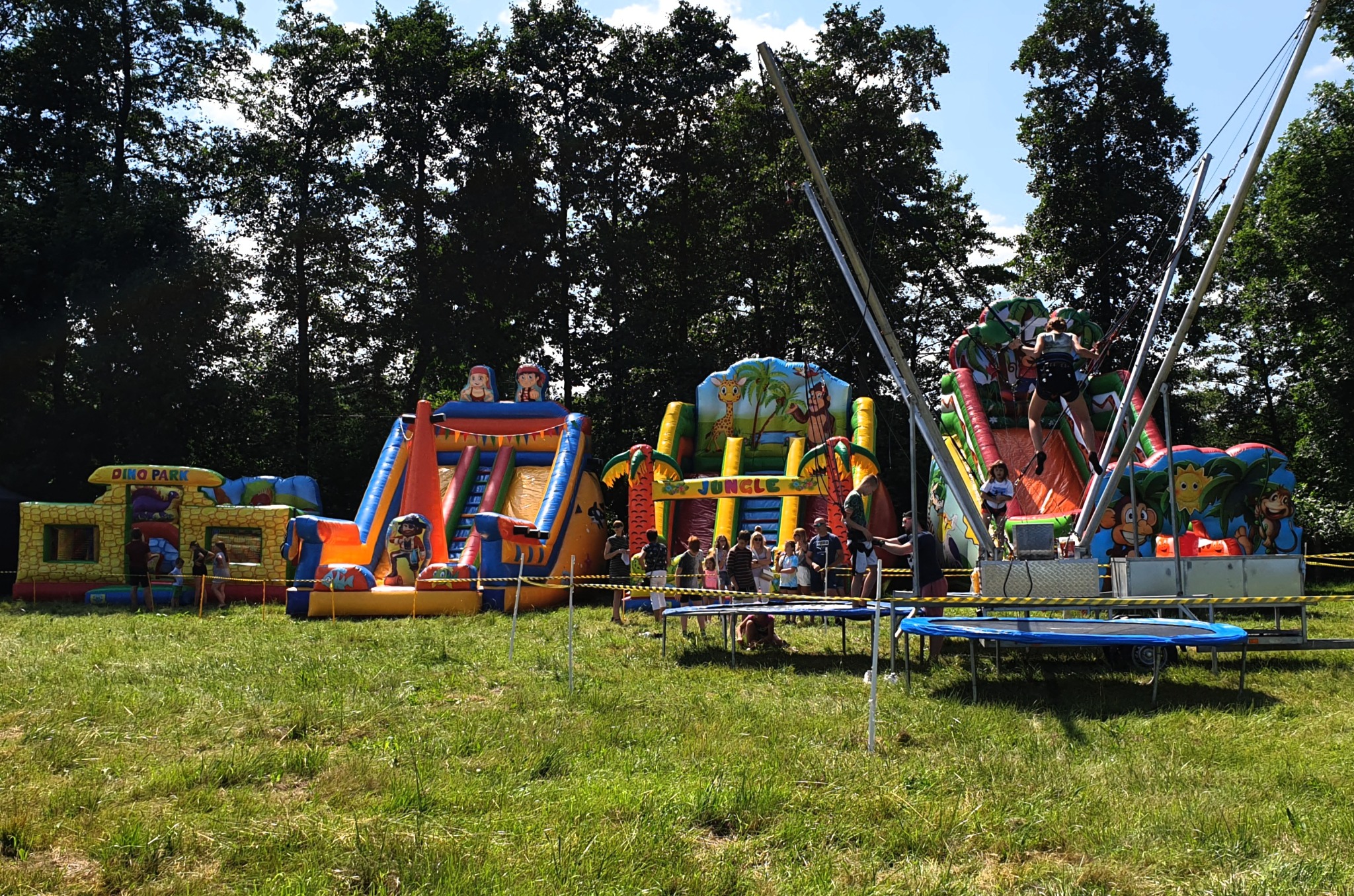 Kolorowe, dmuchane konstrukcje dla dzieci na zielonej trawie, w tle drzewa, widoczni ludzie korzystający z atrakcji.