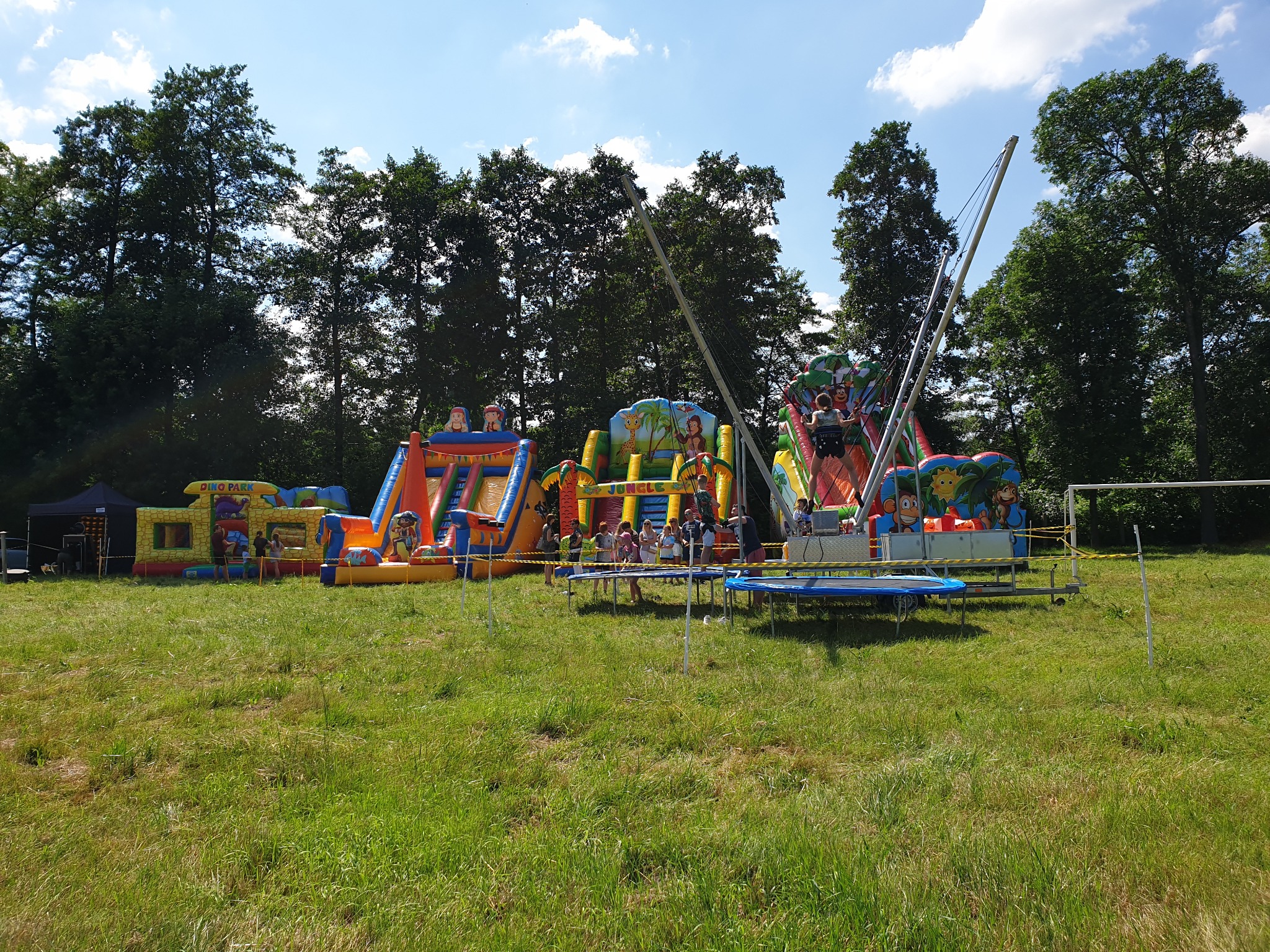 Rozległy park rozrywki na trawiastym terenie z dmuchanymi zamkami, zjeżdżalniami i trampolinami bungee dla dzieci, otoczony drzewami i błękitnym niebem.