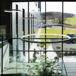 Wnętrze nowoczesnego budynku z przeszkloną ścianą, widok na zewnątrz z trawnikiem, drzewami i zaparkowanym samochodem, oświetlenie w postaci okrągłych lamp, szklane balustrady schodów i roślina...