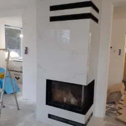 Obudowa kominka wykończona białymi płytami imitującymi marmur, z czarnymi akcentami poziomymi, w trakcie prac wykończeniowych, widoczny drabina i materiały budowlane.