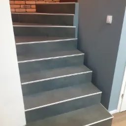 Schody wykończone szarą płytką z białymi listwami, ściana z cegły dekoracyjnej, szara ściana z włącznikiem światła, podłoga drewniana.