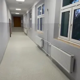 Wnętrze długiego, pustego korytarza z jasną podłogą i szarymi ścianami, widoczne białe grzejniki pod oknami i drzwi na końcu korytarza, oświetlenie sufitowe.