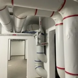 Białe rury instalacyjne pod sufitem w pomieszczeniu technicznym, widoczne zawory i izolacja, w tle otwarte przejście do kolejnego pomieszczenia.