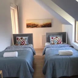 Pokój na poddaszu z dwoma pojedynczymi łóżkami, szarą pościelą, wzorzystymi poduszkami i ręcznikami na każdym łóżku, obraz z motywem plaży na ścianie, drewniana podłoga.