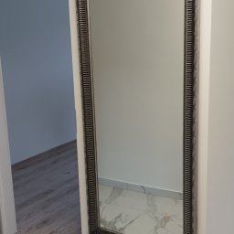 IGOTECH Sp. z o.o. - Wnętrze po remoncie: duże lustro w ozdobnej ramie zawieszone na białej ścianie, zabezpieczone niebieską taśmą malarską przy podłodze i na krawędzi ściany, widoczna marmurowa podłoga i fragment...