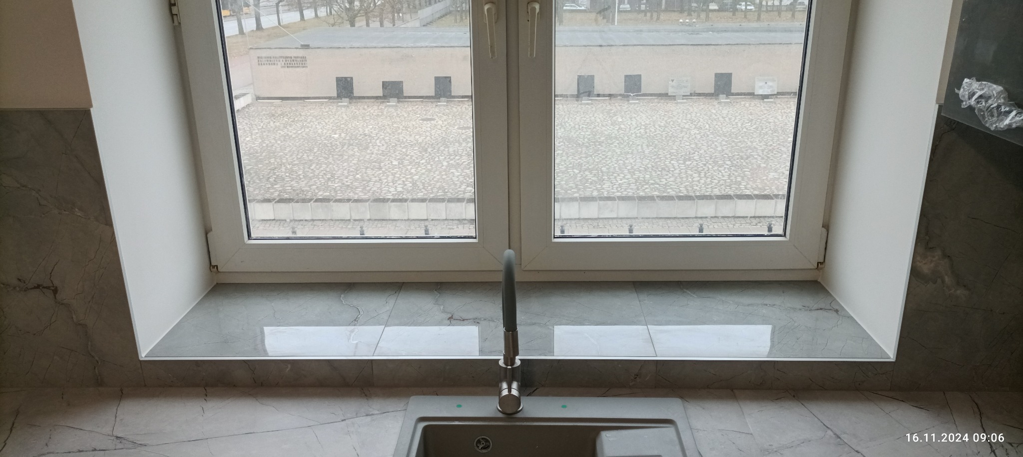 Szary zlewozmywak z baterią na tle okna z widokiem na cmentarz, wykończony płytkami imitującymi marmur.
