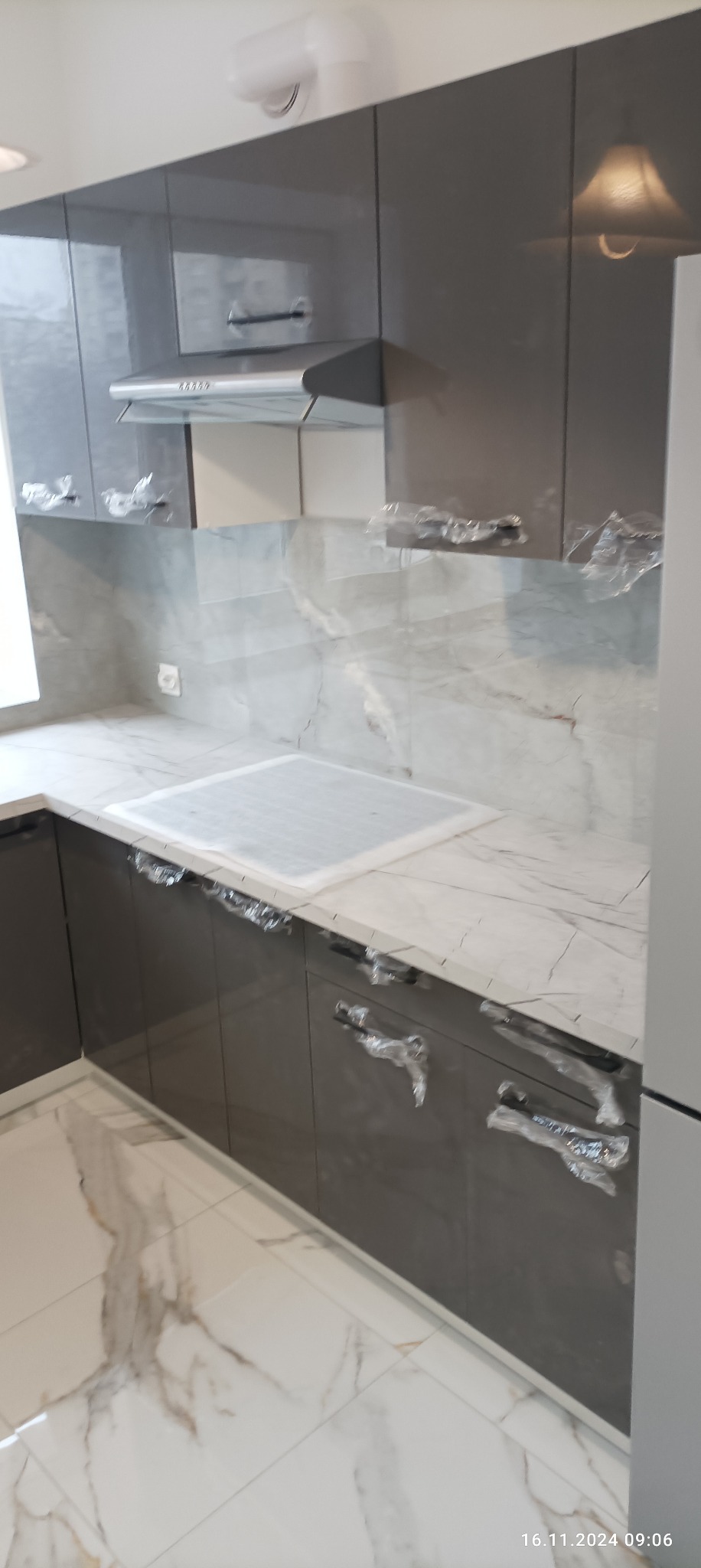 Nowoczesna kuchnia z szarymi, lakierowanymi szafkami, marmurowym blatem i backsplashiem, z folią ochronną na uchwytach i płycie grzewczej, świeżo po montażu.