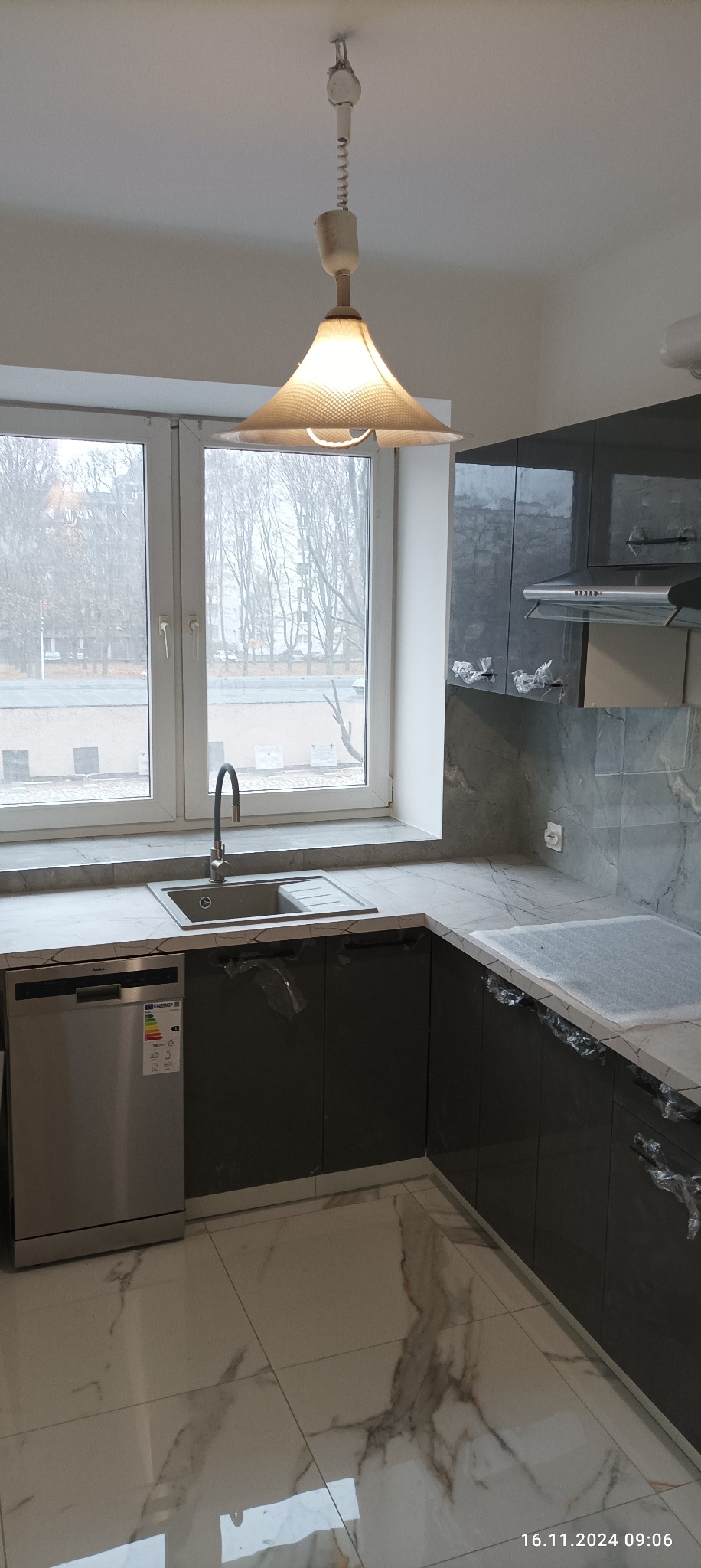 Szara, błyszcząca kuchnia z marmurowym blatem i backsplashiem, z widocznym zlewem, baterią i zmywarką, częściowo zabezpieczona folią ochronną, oświetlona lampą z wiklinowym abażurem.