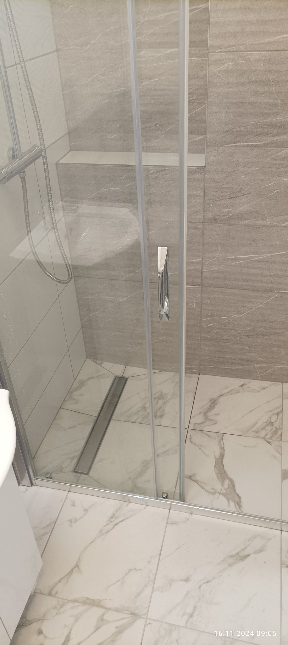 Szklana kabina prysznicowa z odpływem liniowym w nowoczesnej łazience wykończonej płytkami imitującymi marmur.