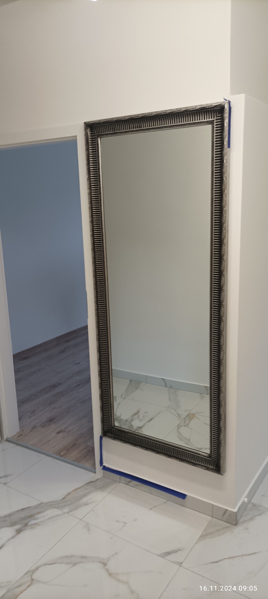 Wnętrze po remoncie: duże lustro w ozdobnej ramie zawieszone na białej ścianie, zabezpieczone niebieską taśmą malarską przy podłodze i na krawędzi ściany, widoczna marmurowa podłoga i fragment...