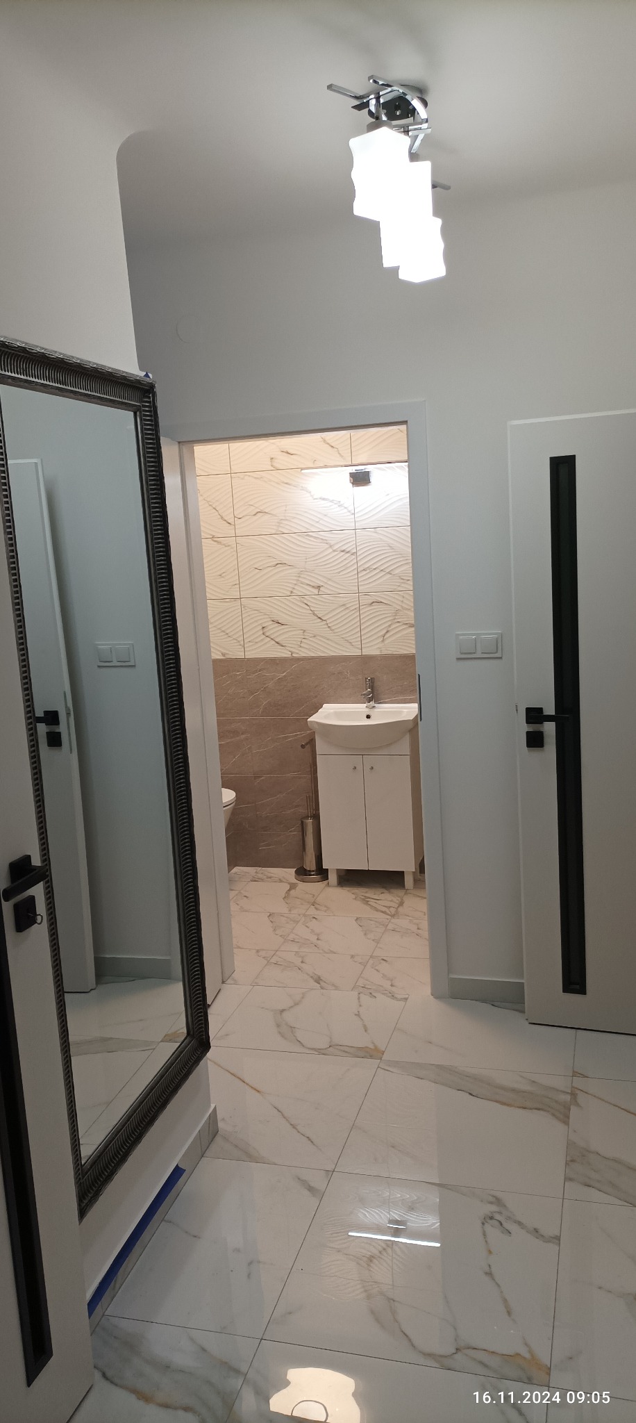 Wykończona łazienka z marmurową podłogą, beżowymi płytkami imitującymi fale na ścianach, białą szafką pod umywalką i ozdobnym lustrem w ciemnej ramie, widok z korytarza.