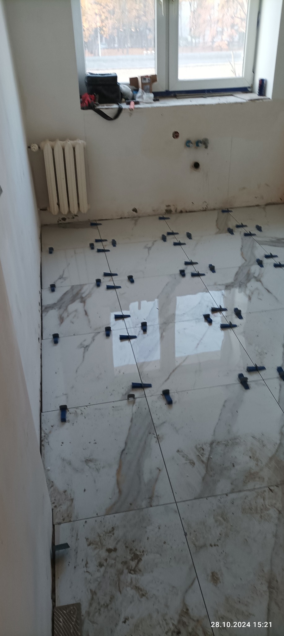 Proces układania płytek podłogowych imitujących marmur, z użyciem systemu poziomowania płytek, widoczne krzyżyki dystansowe i sznurki wyznaczające linie, w tle okno i grzejnik.