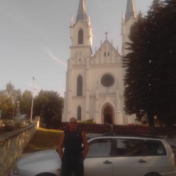 Usługi budowlane - Mężczyzna w okularach przeciwsłonecznych stoi przed srebrnym kombi z drabiną na dachu, na tle białego kościoła z dwiema wieżami.