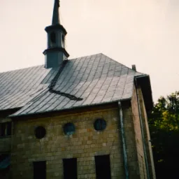 Kamienny budynek z szarym dachem pokrytym dachówką, widoczny fragment drabiny dachowej, okrągłe okna i rynny.