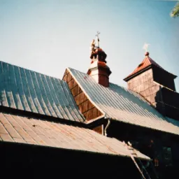 Stary, drewniany kościół z blachy falistej na dachu i drewnianą wieżą z krzyżem, z pracownikiem na drabinie przy rynnie.