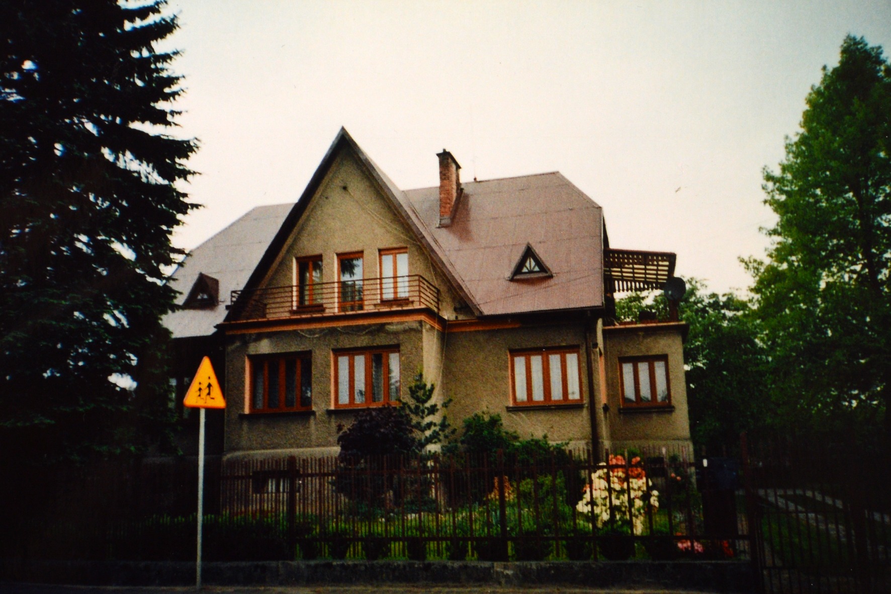 Duży, dwupiętrowy dom z brązowym dachem i kremową elewacją, widoczny zza znaku drogowego ostrzegającego przed przejściem dla pieszych, otoczony zielenią drzew i krzewów.