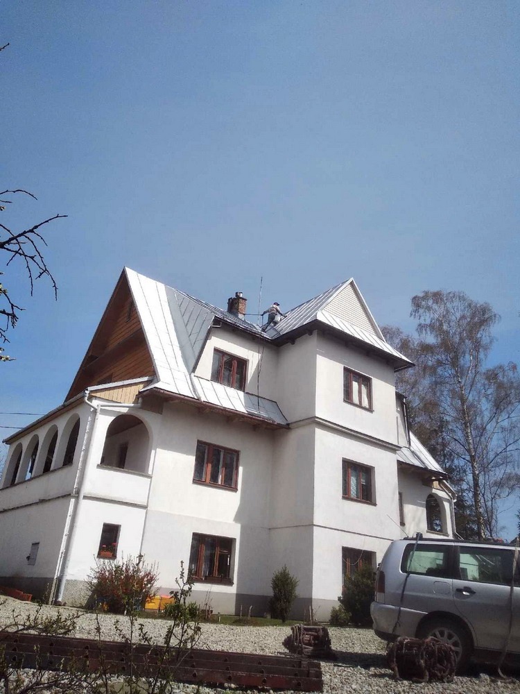 Dwupiętrowy, biały dom z szarym, metalowym dachem widziany z dołu. Na dachu widoczna sylwetka osoby pracującej w jasnych ubraniach. Na pierwszym planie zaparkowany srebrny samochód i fragmenty...