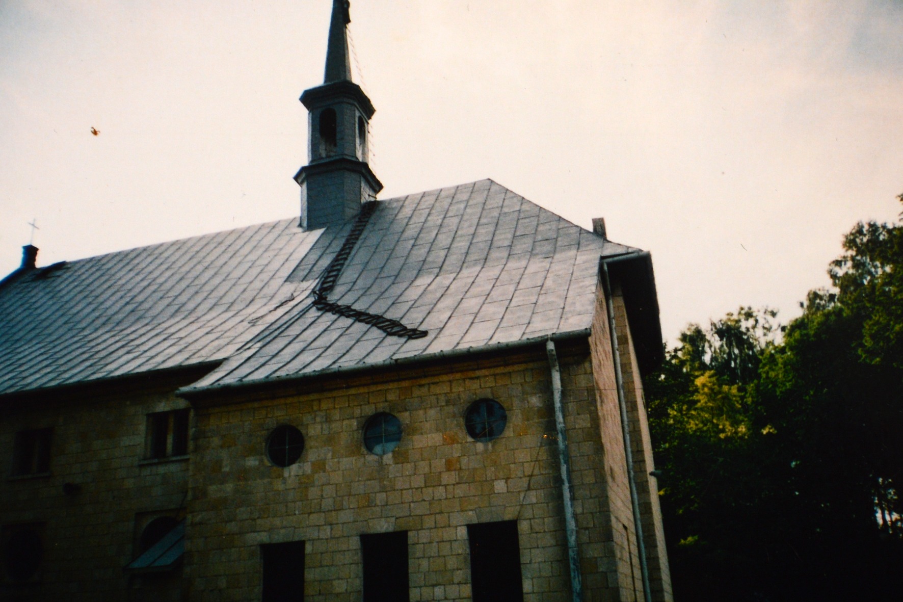 Kamienny budynek z szarym dachem pokrytym dachówką, widoczny fragment drabiny dachowej, okrągłe okna i rynny.