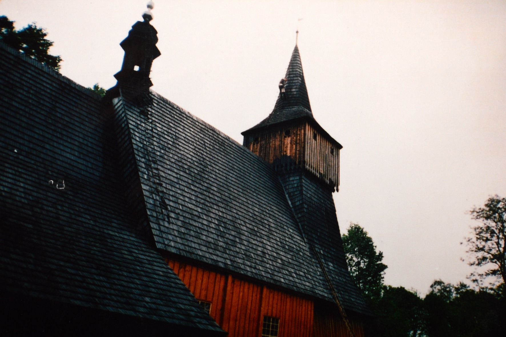 Fragment zabytkowego, drewnianego kościoła z ciemnym, gontowym dachem i wysoką wieżą, sfotografowany pod kątem z dołu w pochmurny dzień.
