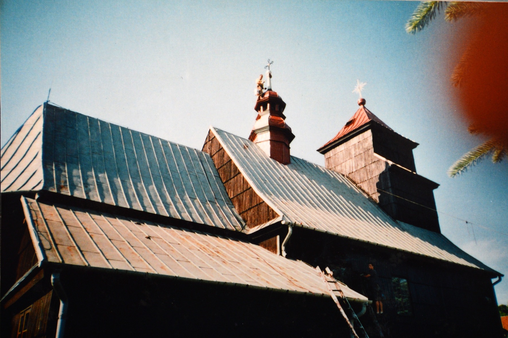 Stary, drewniany kościół z blachy falistej na dachu i drewnianą wieżą z krzyżem, z pracownikiem na drabinie przy rynnie.