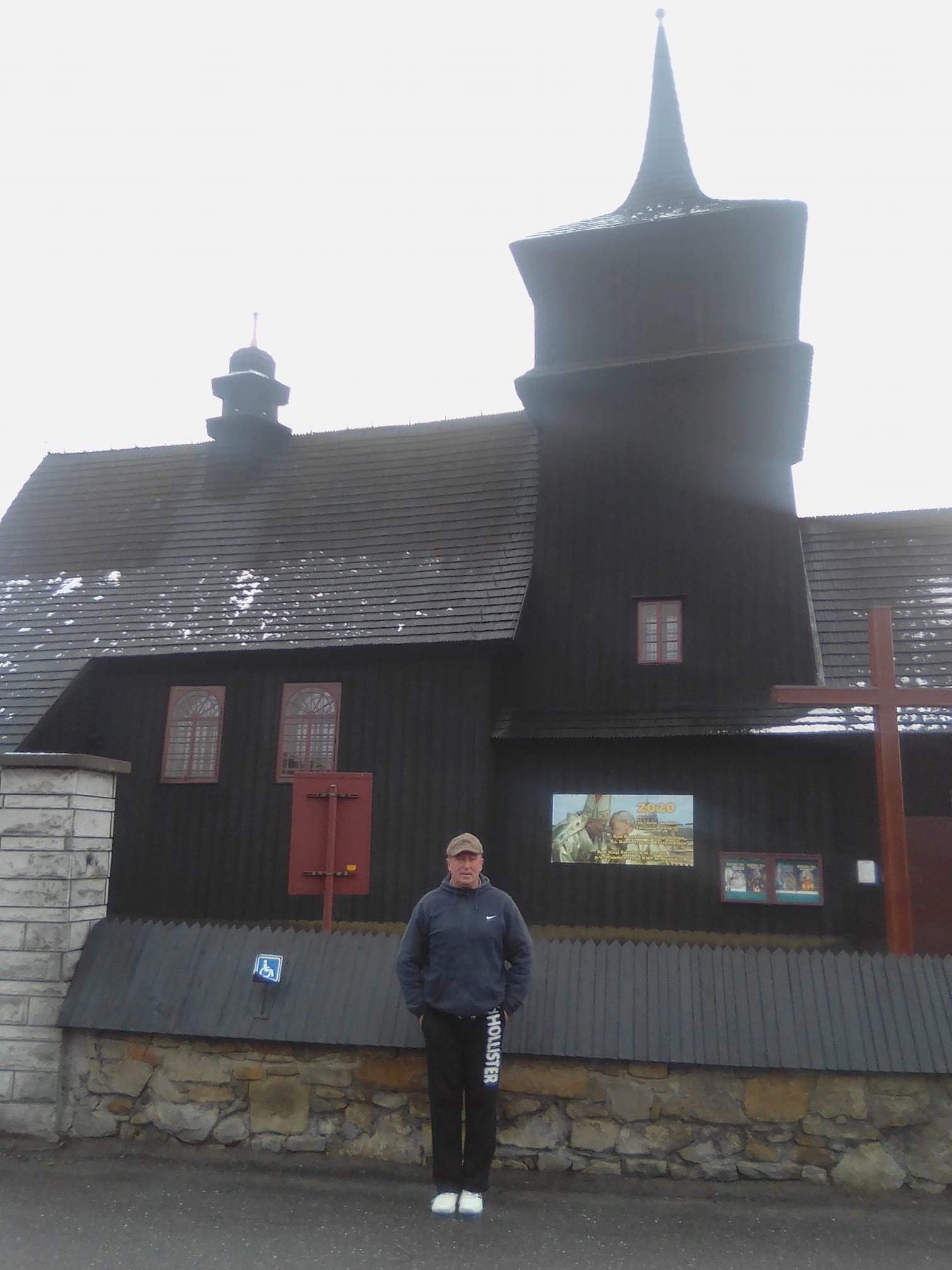 Mężczyzna w sportowym stroju stoi na tle ciemnego, drewnianego kościoła z wieżą zwieńczoną spiczastym dachem, na dachu widoczne ślady śniegu, na ścianie budynku wisi plakat.