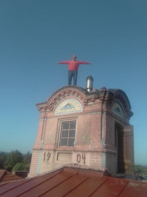 Mężczyzna w czerwonej koszuli stoi na szczycie wieżyczki dachowej z datą 1904, rozkładając ręce na tle błękitnego nieba.