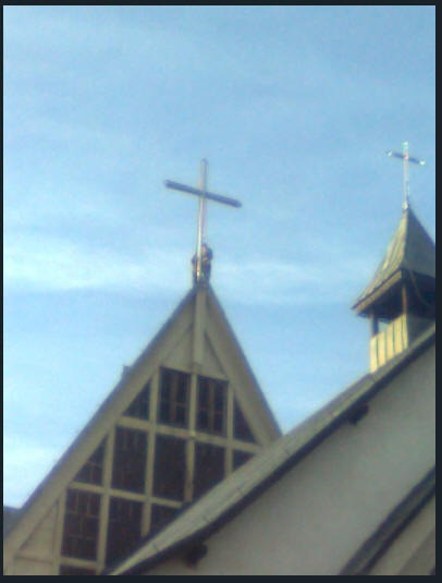 Fragment dachu kościoła z krzyżem, widoczne okna na szczycie i fragment wieżyczki z drugim krzyżem na tle błękitnego nieba.