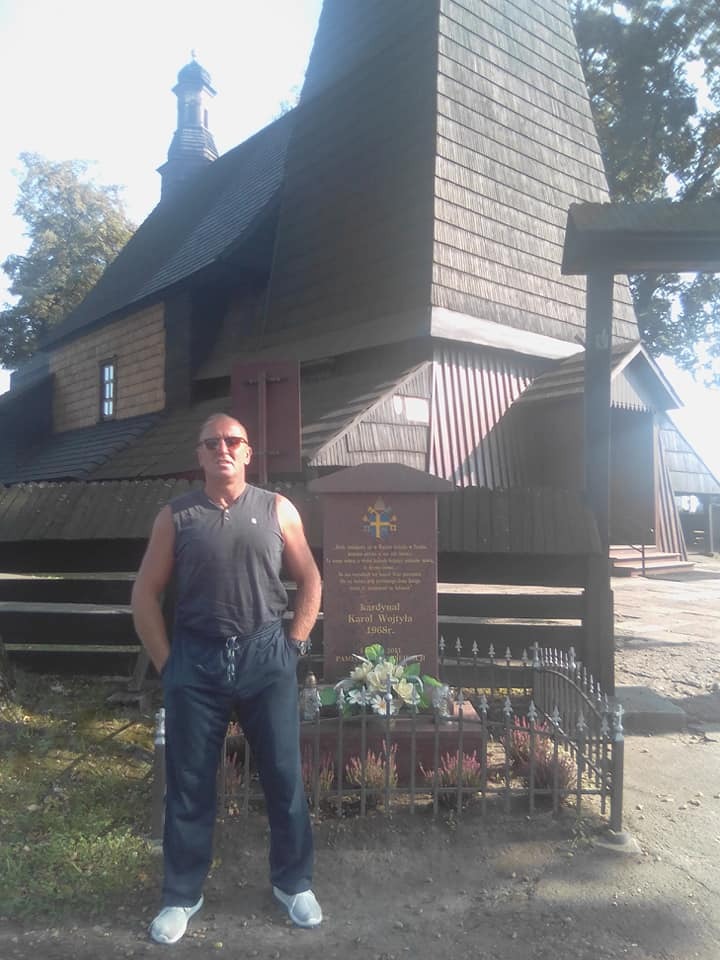 Mężczyzna w sportowym stroju stoi na tle drewnianego kościoła z charakterystyczną, wysoką wieżą i pomnika kardynała Karola Wojtyły.
