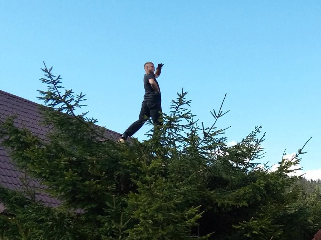 Mężczyzna w czarnym stroju stoi na dachu za zielonymi drzewami, machając ręką na tle błękitnego nieba. Nietypowy kadr z dołu.