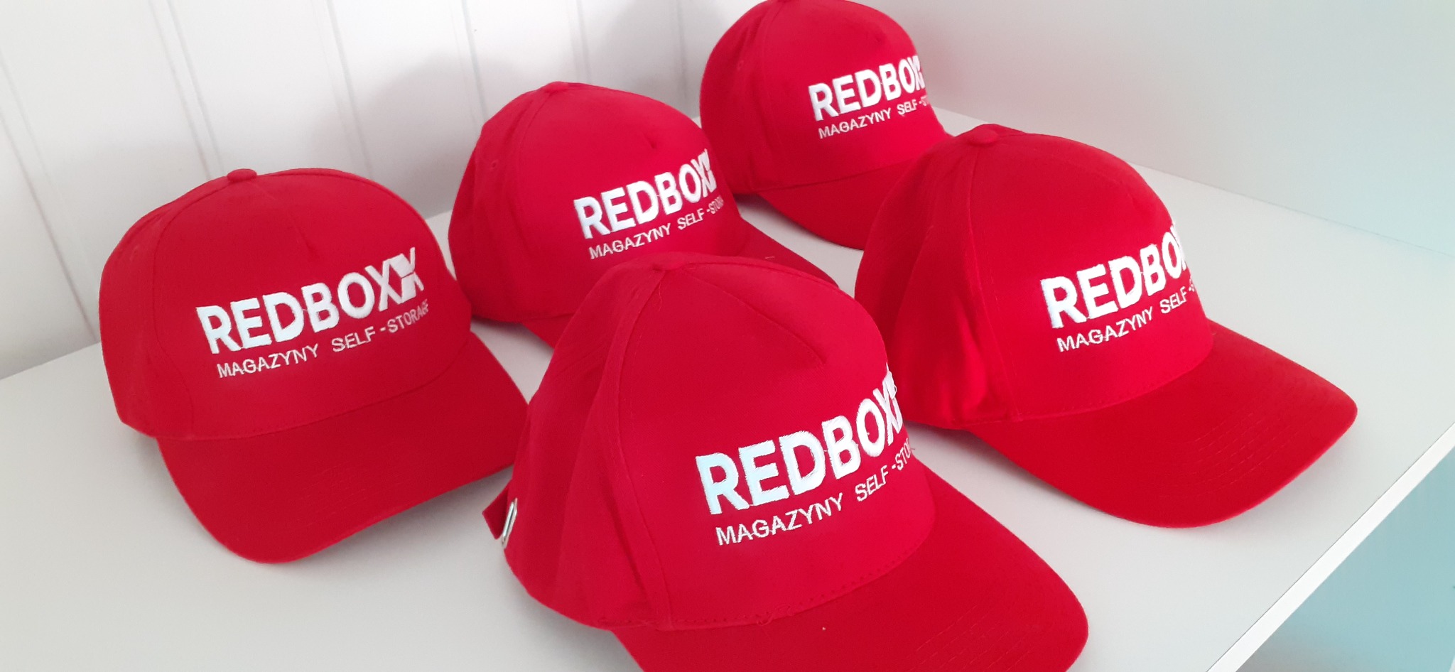 Czerwone czapki z daszkiem z białym nadrukiem 'REDBOXX Magazyny Self-Storage' ułożone na białym tle.