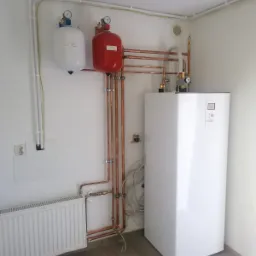 Biała jednostka wewnętrzna z czerwonym i białym zbiornikiem ciśnieniowym, połączona miedzianymi rurami, zamontowana w rogu pomieszczenia z grzejnikiem.