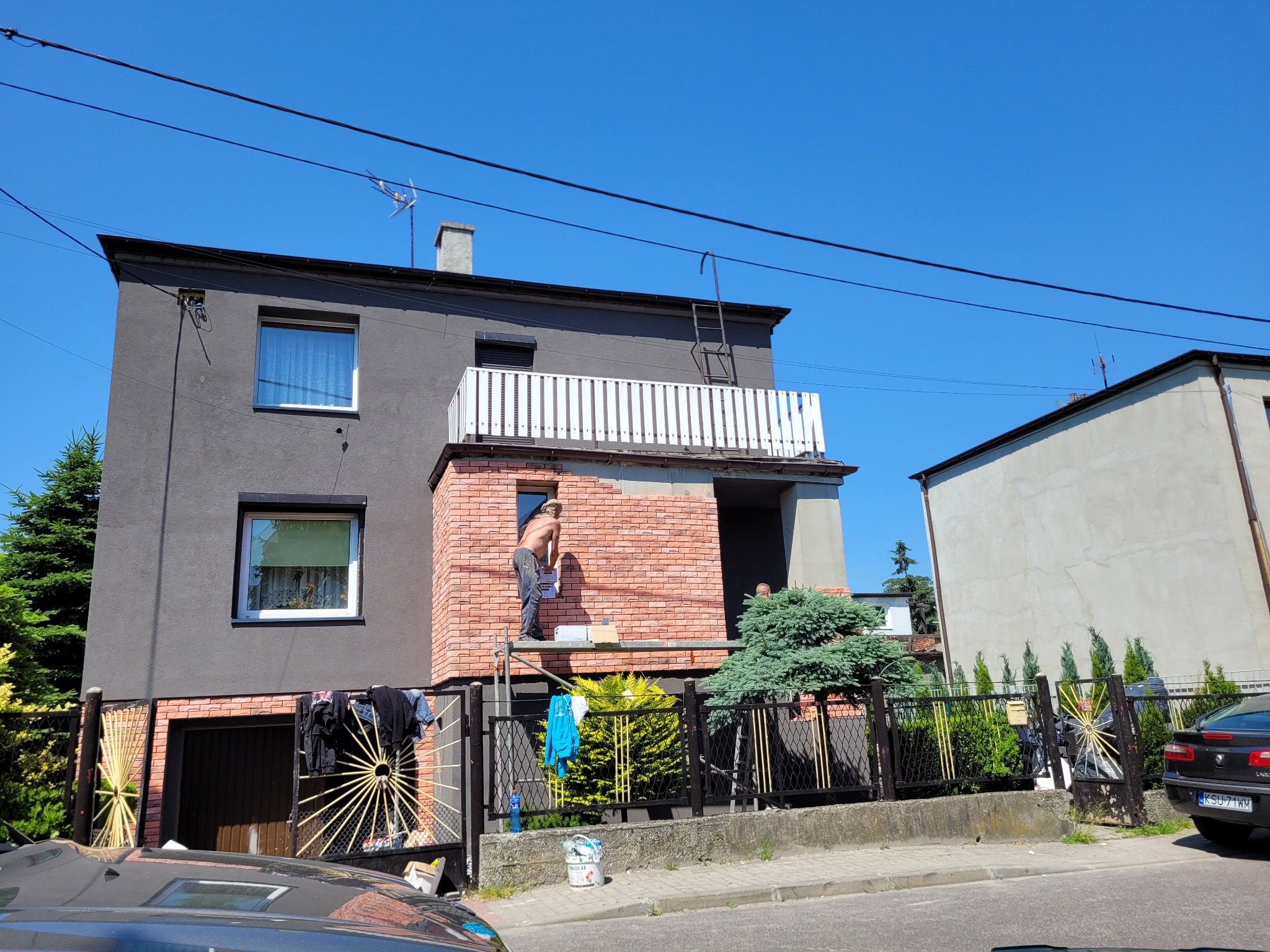 Mężczyzna bez koszulki na rusztowaniu, nakładający zaprawę na ceglaną elewację domu jednorodzinnego w słoneczny dzień. Widoczny fragment elewacji pomalowany na szaro, a w tle błękitne niebo.