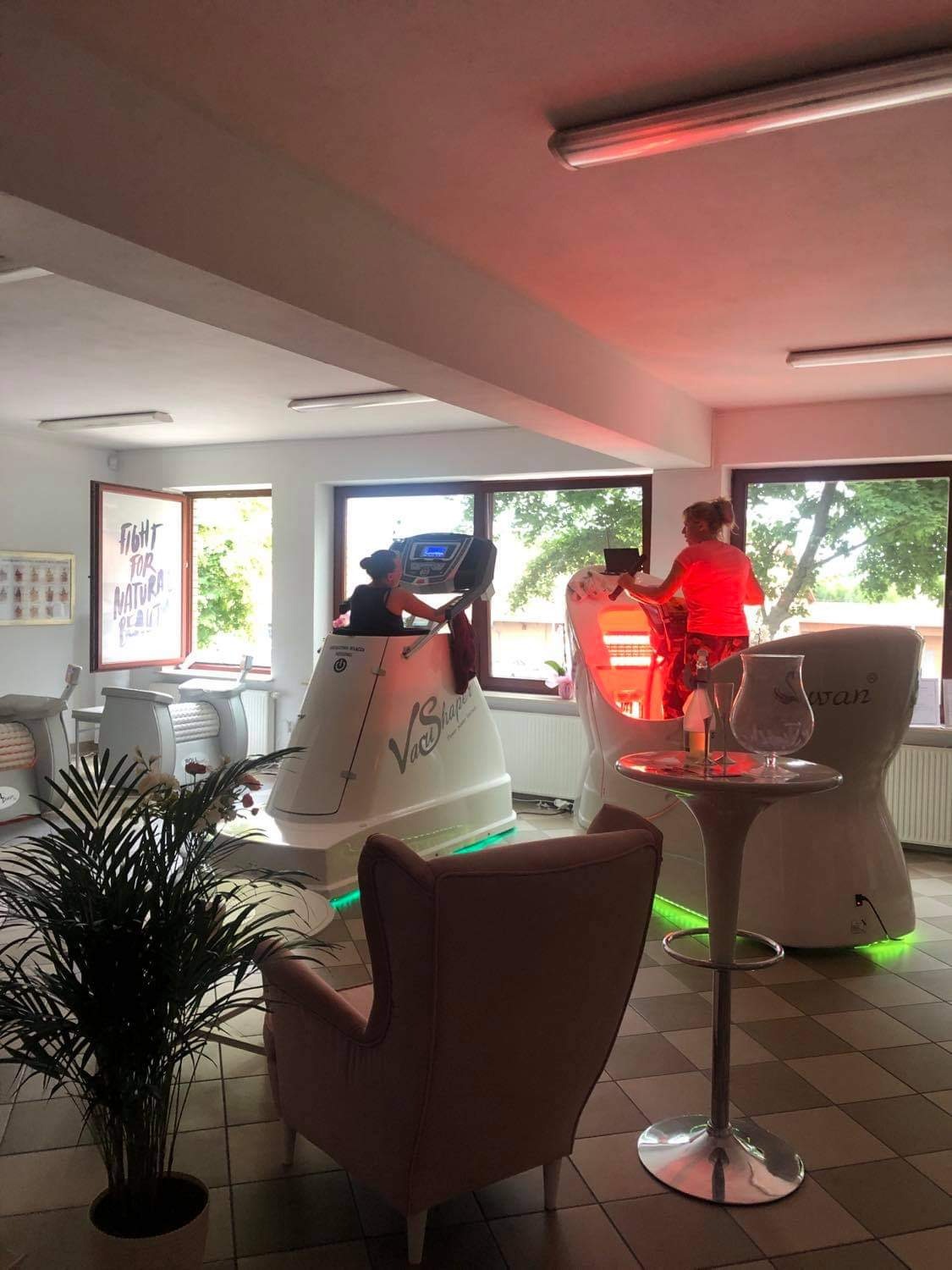 Wnętrze gabinetu z urządzeniami do modelowania sylwetki: osoba ćwiczy na bieżni w kapsule VacuShape, druga osoba obsługuje urządzenie SwanShape z czerwonym światłem. W tle okno z napisem 'Fight for...