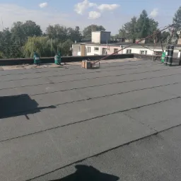 Wykończony, płaski dach pokryty papą termozgrzewalną z widocznymi butlami gazowymi, narzędziami i dźwigiem do transportu materiałów, w tle zabudowa miejska i drzewa.